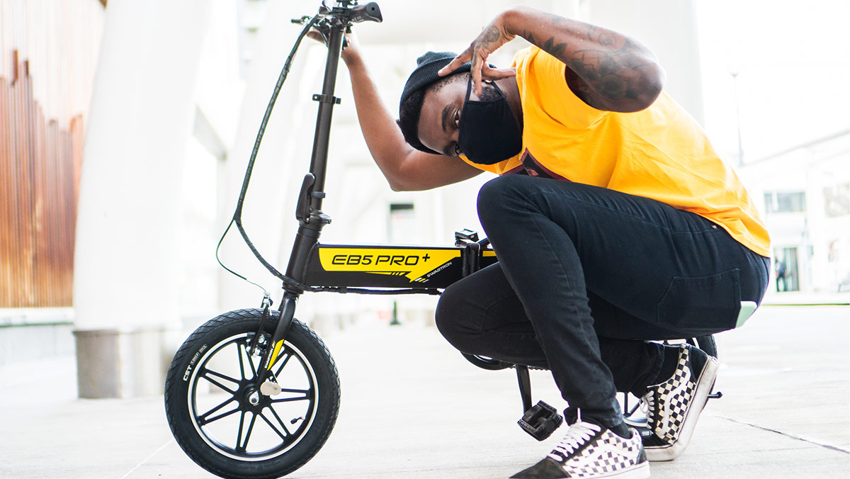 Rossini wearing black pants and yellow shirt squatting next to eb5 pro plus ebike in denver downtown.